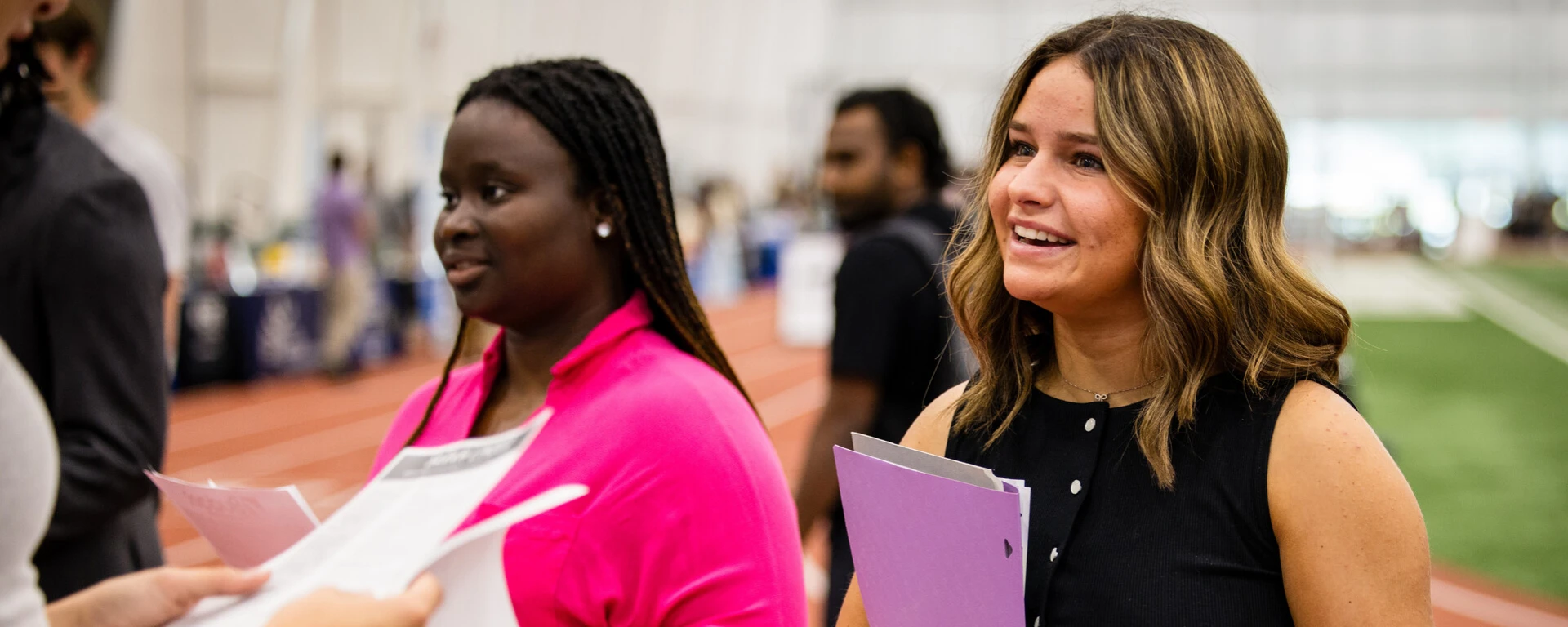Two women smiling while holding papers and a purple folder at career and internship event with other people in the background at Northwest Missouri State University