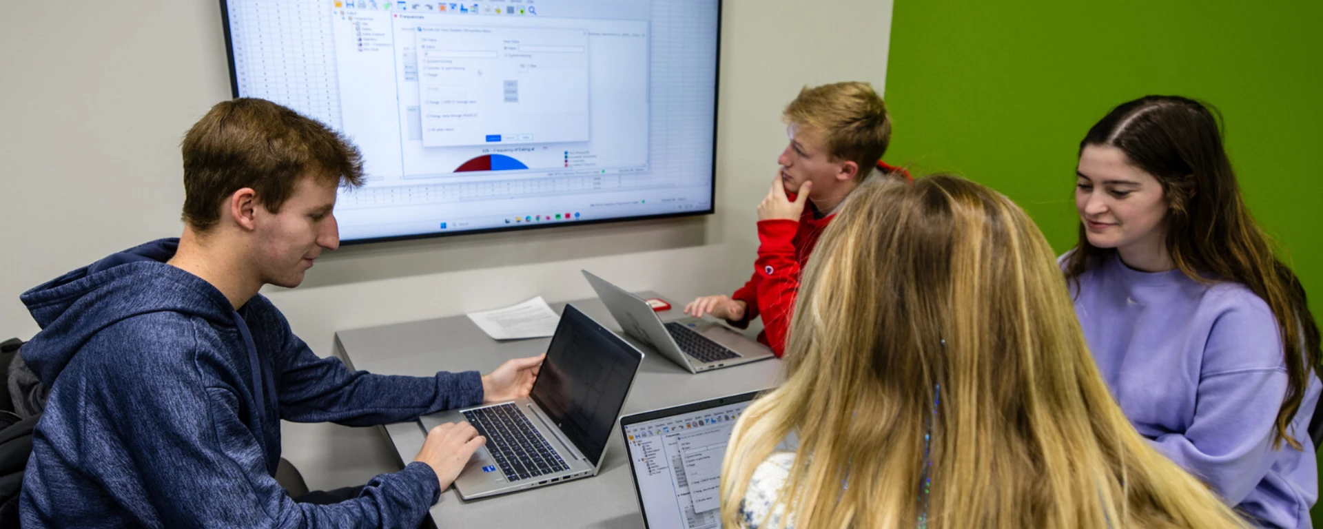 Three people working together at a desk with laptops, looking at a large monitor displaying a spreadsheet with data and a dialog box open, pointing at the screen to discuss the information displayed at Northwest Missouri State University