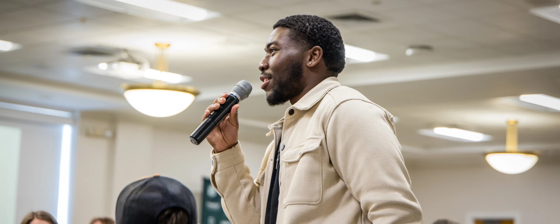 A man with a beard wearing a beige jacket speaks into a microphone while addressing a seated audience in an indoor venue with fluorescent ceiling lights at Northwest Missouri State University