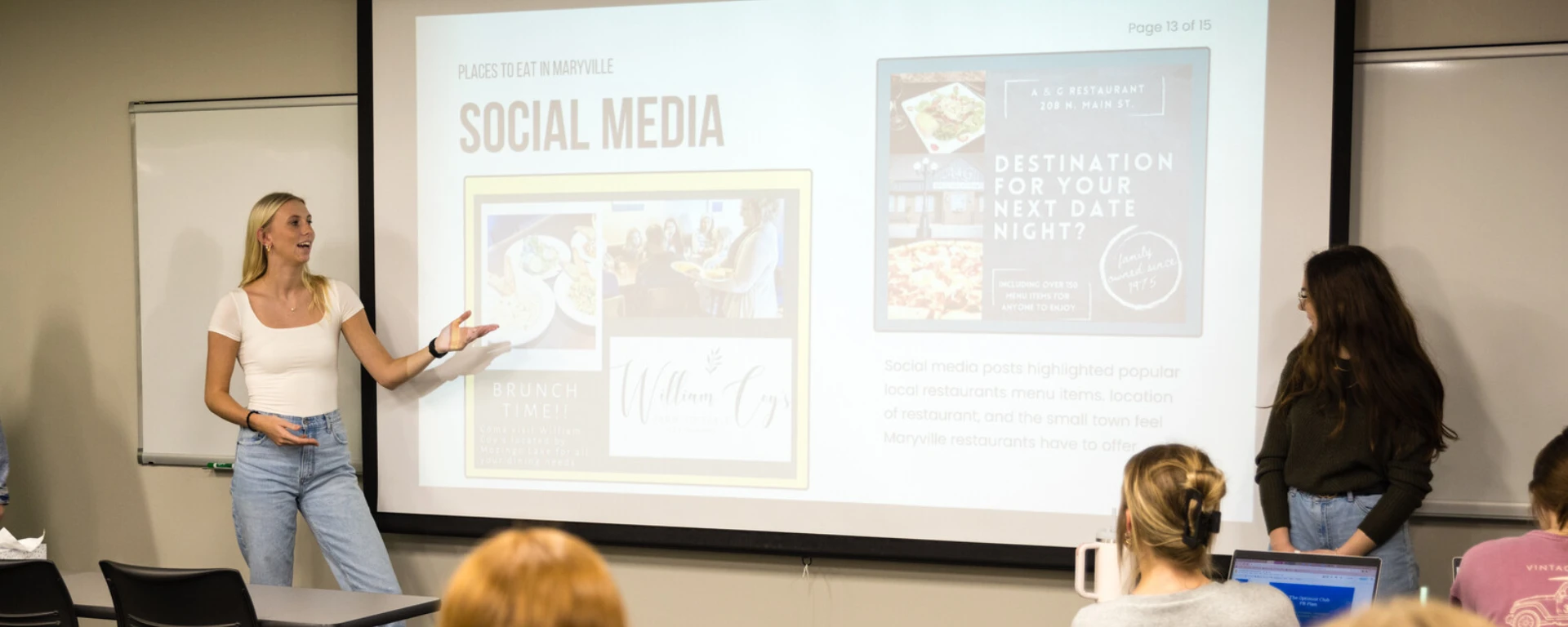 A female presenter gestures toward a projector screen displaying social media content while teaching a class of students seated at desks with laptops in a classroom setting at Northwest Missouri State University