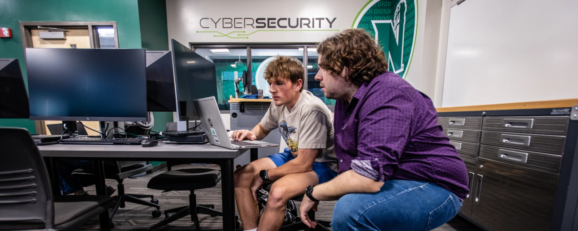 An adult instructor in a purple shirt crouches beside a young student sitting at a desk with a laptop in a cybersecurity lab, with a CYBERSECURITY sign visible on the wall behind them at Northwest Missouri State University
