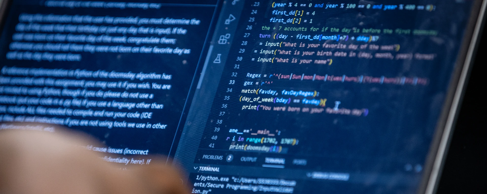 A close-up view of a computer screen displaying lines of blue-colored source code and programming syntax with a blurred hand pointing at or interacting with the monitor at Northwest Missouri State University