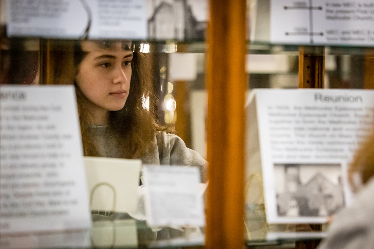 Northwest student Nora Crowley is reflected in a display case at the Nodaway County Historical Society Museum as she organized pieces of an exhibit detailing religious history in the region. (Photo by Lilly Cook/Northwest Missouri State University)